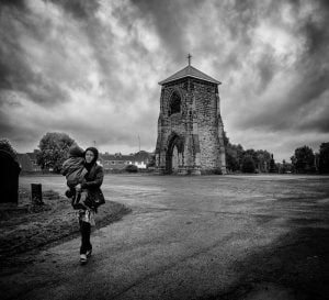 A woman walking with a baby in front of a church tower.