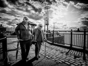 A monochrome image capturing an elderly couple strolling along the coastal sidewalk.