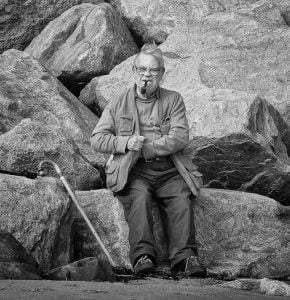 Street photography of an elderly man sitting on rocks with a cane.