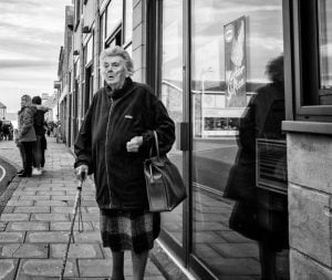 Street photography of an elderly woman gracefully strolling down the street, aided by a cane.