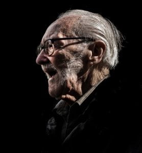 An elderly man wearing glasses captured in a street photography style against a black background.