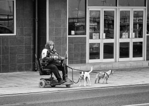 Street photography of a wheelchair-bound woman walking her two dogs on a leash.